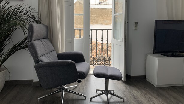 Un salon moderne avec un canapé gris, un petit pouf, un téléviseur posé sur un meuble blanc et une grande fenêtre donnant sur un immeuble.