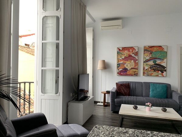 Una sala moderna con un sofá gris, una mesa de centro y un televisor sobre un mueble blanco. Hay dos cuadros de peces en la pared y una ventana con cortinas.