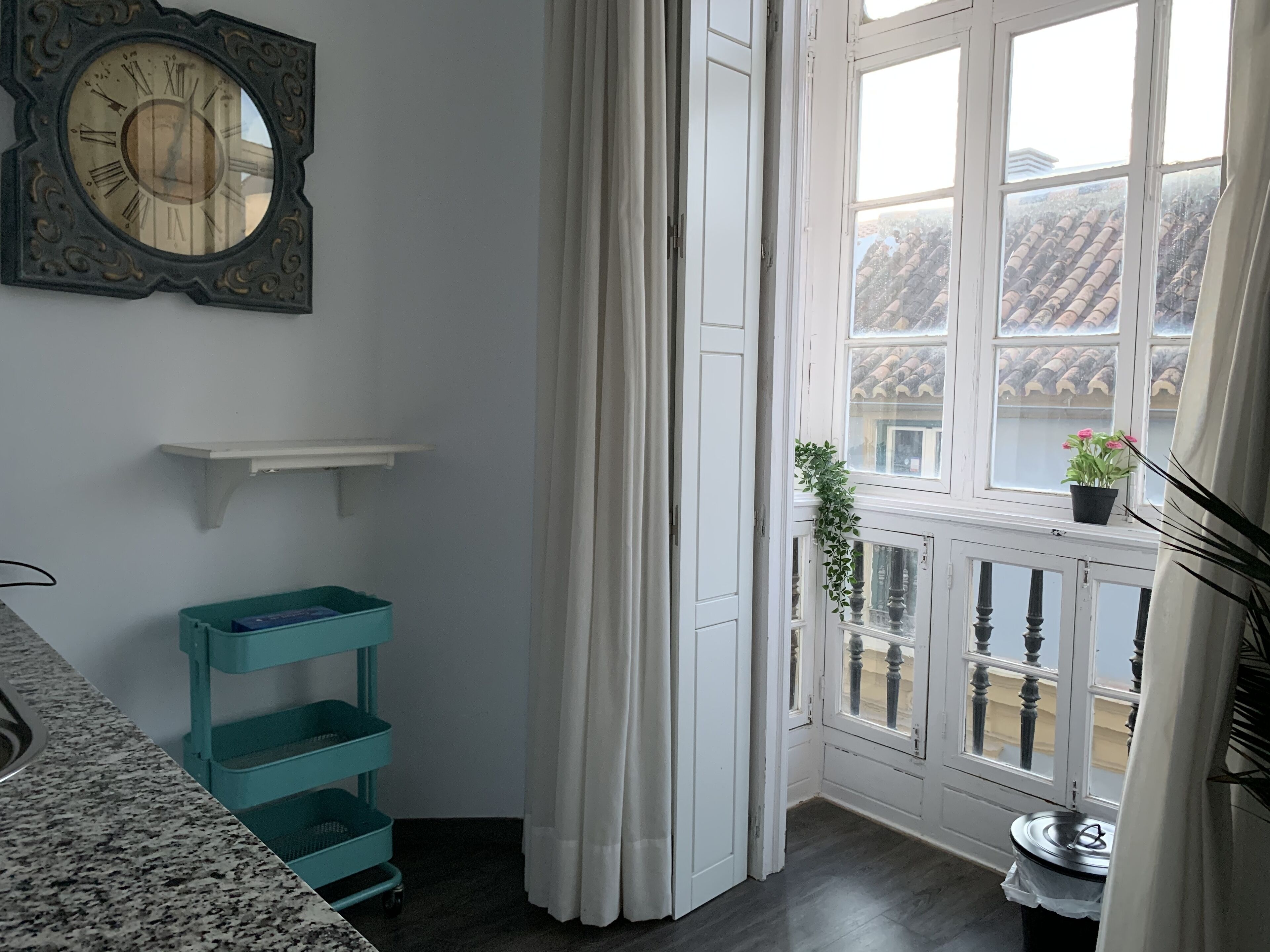 A room with a large wall clock, a window with a view of a building, a potted plant on the windowsill, and a teal storage unit.