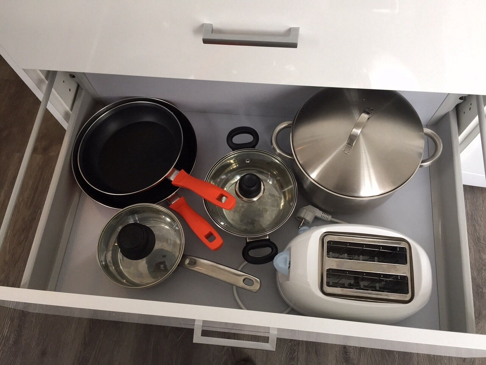 A kitchen drawer containing a black frying pan, a stainless steel pot, a saucepan, a toaster, and a set of lids.