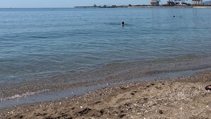 Une plage à l’eau claire, une personne qui nage et, au loin, des structures industrielles.