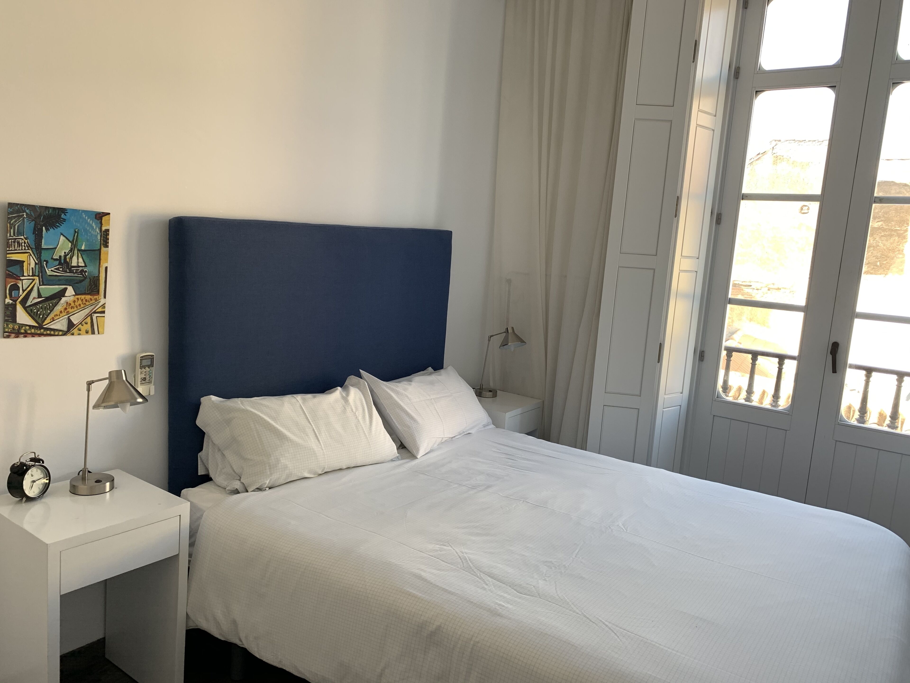 Un dormitorio con una cama, mesitas de noche blancas, una lámpara, un reloj y un cuadro en la pared.