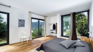 Un dormitorio moderno con una cama grande, piso de madera, vista a zonas verdes y montañas, y una puerta corrediza de vidrio.