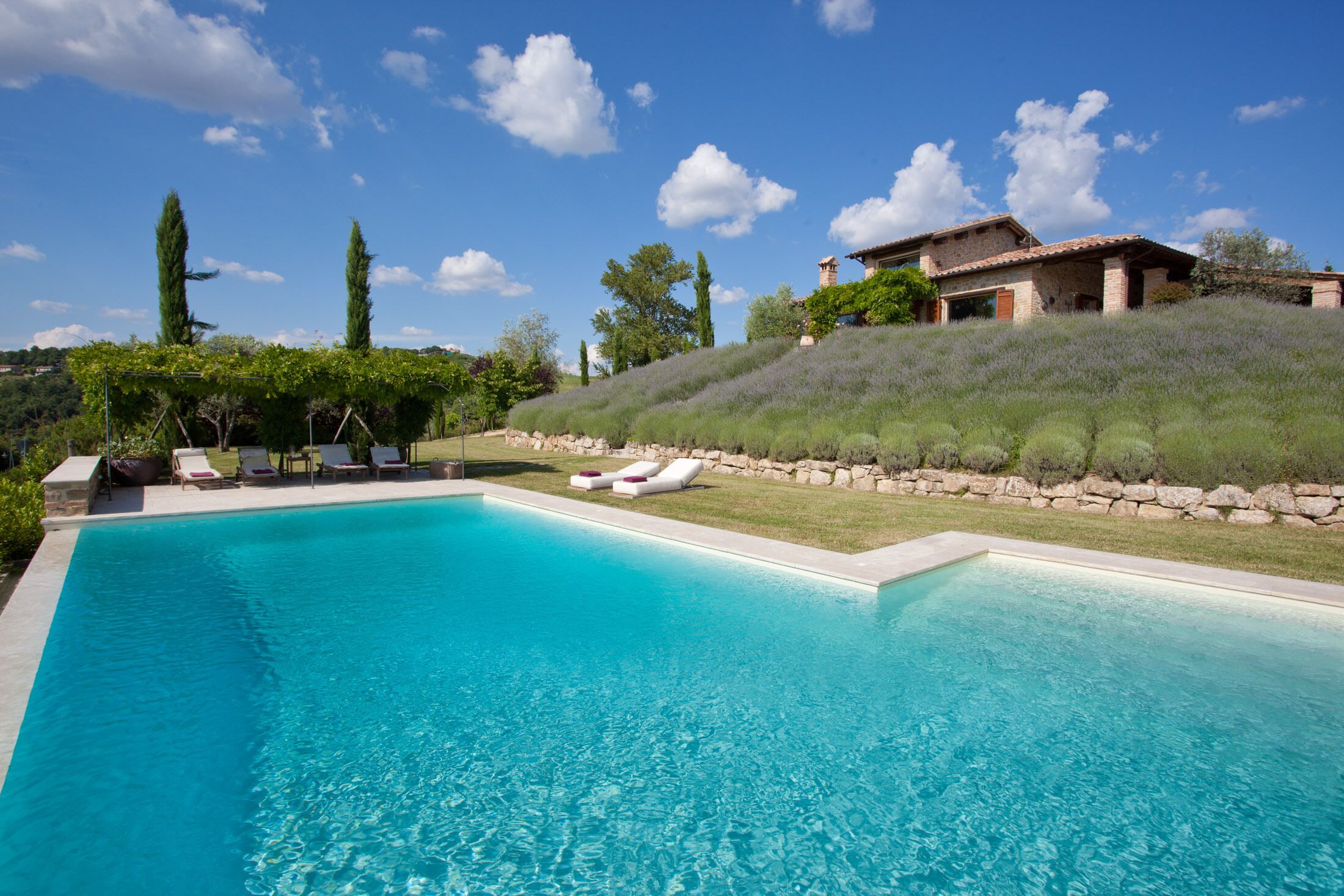 Ein rechteckiger Swimmingpool mit klarem, blauem Wasser, umgeben von einer Steinmauer und einer Rasenfläche mit Liegestühlen.