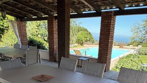 An outdoor patio with a long table and chairs, a brick pillar, and a view of a swimming pool and greenery.