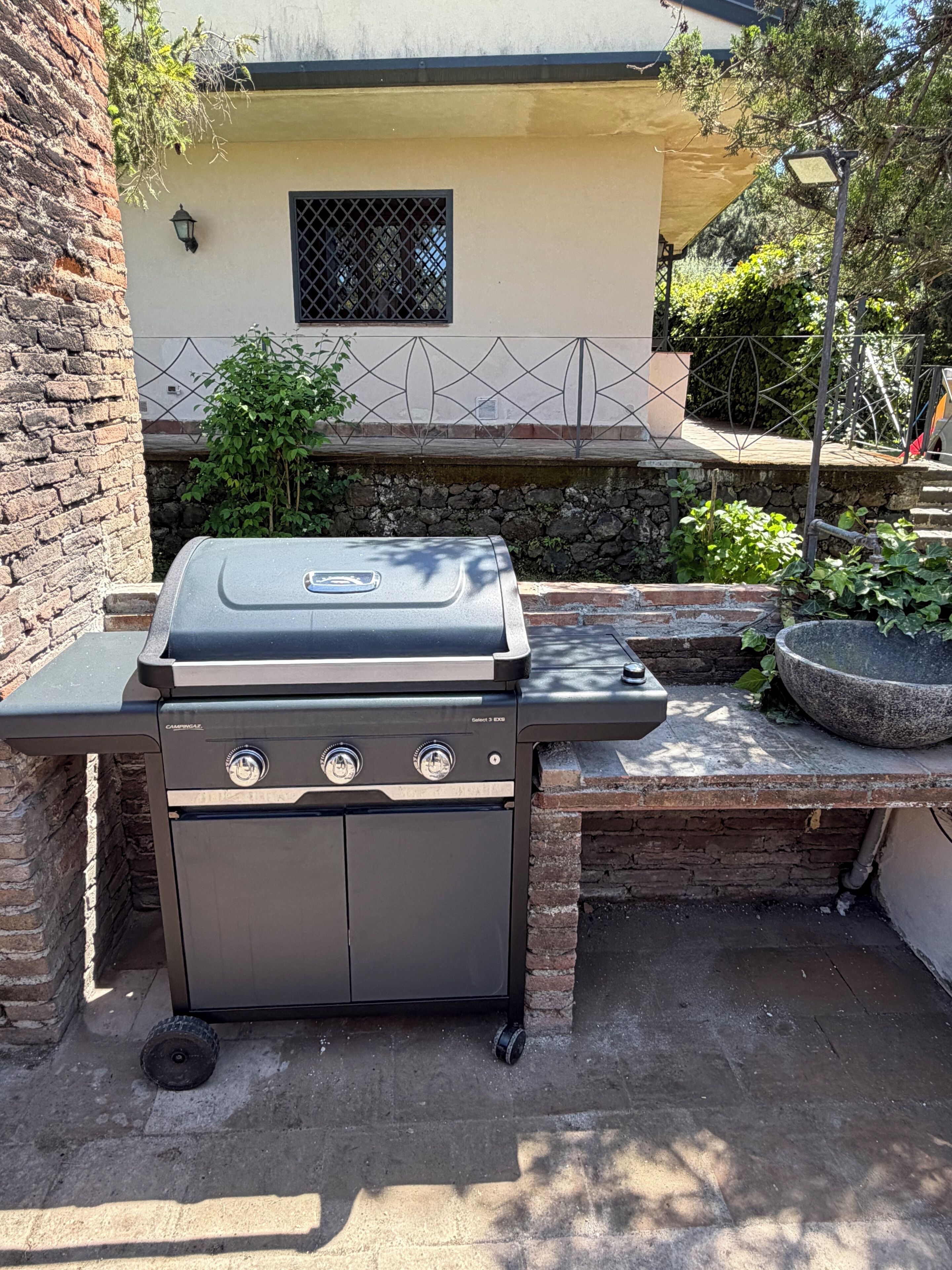 Una parrilla con controles, un muro de piedra y una casa al fondo.