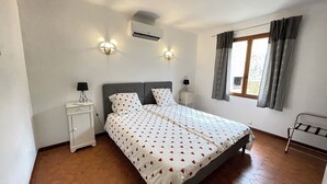 A bedroom with a large bed, two bedside tables with lamps, an air conditioner, and a window with curtains.