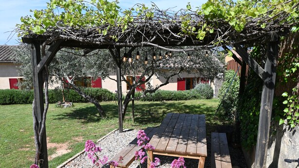 A wooden pergola with a table and benches in a garden area, surrounded by gravel and flowering plants.