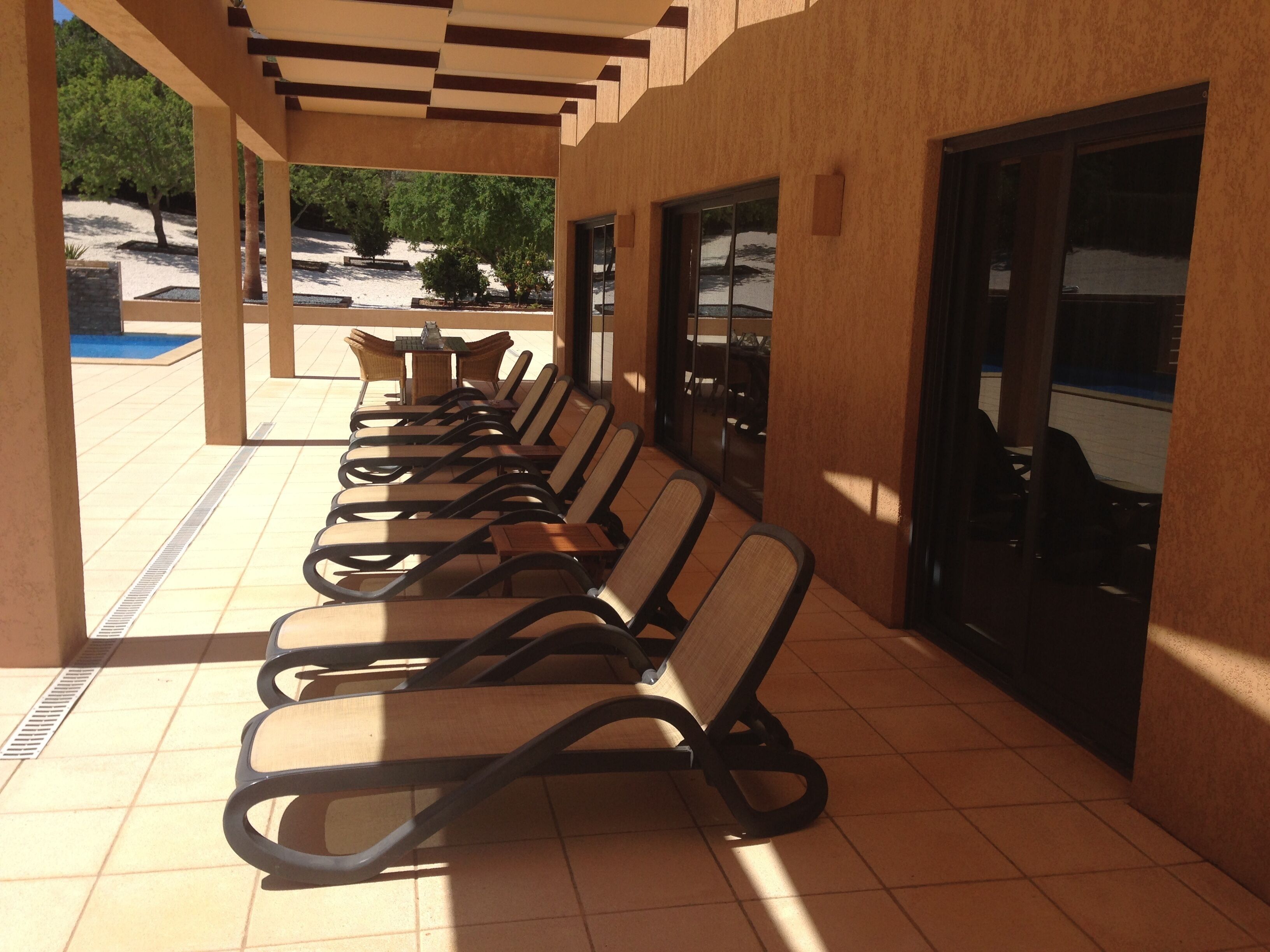 A covered outdoor seating area with rows of lounge chairs and tables, a pool in the background, and a building entrance.