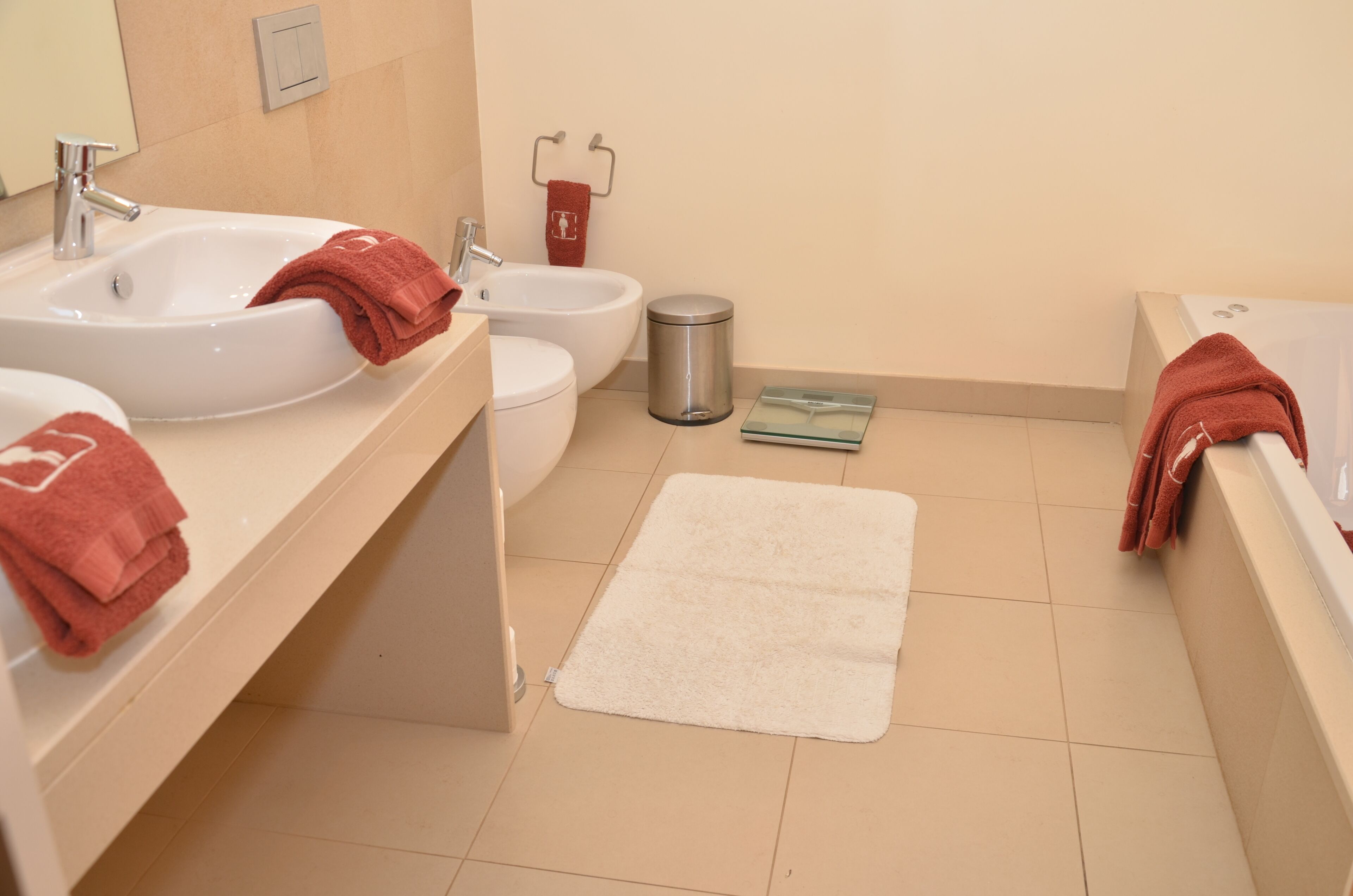 Een badkamer met twee wastafels, een bidet, een toilet, een prullenbak en een witte vloerkleed.