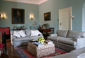 A living room with two sofas, a coffee table, and a red armchair. There are wall-mounted lights and a door. - Historical country house near Taormina & the sea. Stunning view of Mt Etna (Taormina)