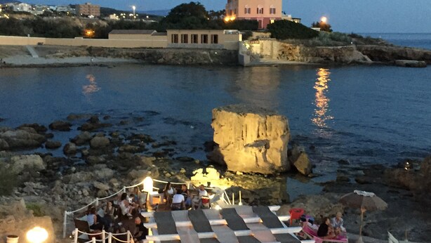 A coastal hotel with a large rock formation in the foreground, a terrace with lounge chairs, and a building with multiple windows in the background. - Studio Cavour (alghero)