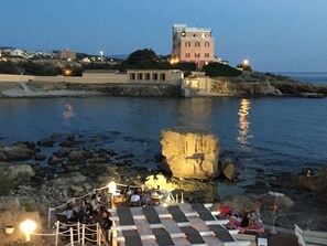 A coastal hotel with a large rock formation in the foreground, a terrace with lounge chairs, and a building with multiple windows in the background. - Studio Cavour (alghero)