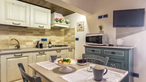 A kitchen with white cabinets, a microwave, a gas stove, and a dining table set with a bowl of fruit and a coffee cup. - Studio Cavour (alghero)