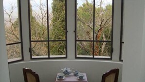 Ein Eckzimmer mit zwei Sesseln und einem Tisch, große Fenster mit Blick auf Bäume und einen klaren Himmel.