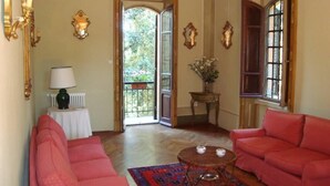Ein Zimmer mit zwei roten Sofas, einem kleinen Tisch mit zwei weißen Gegenständen, einem gemusterten Teppich, einem Heizkörper und einem Fenster mit Blick auf Grünflächen.