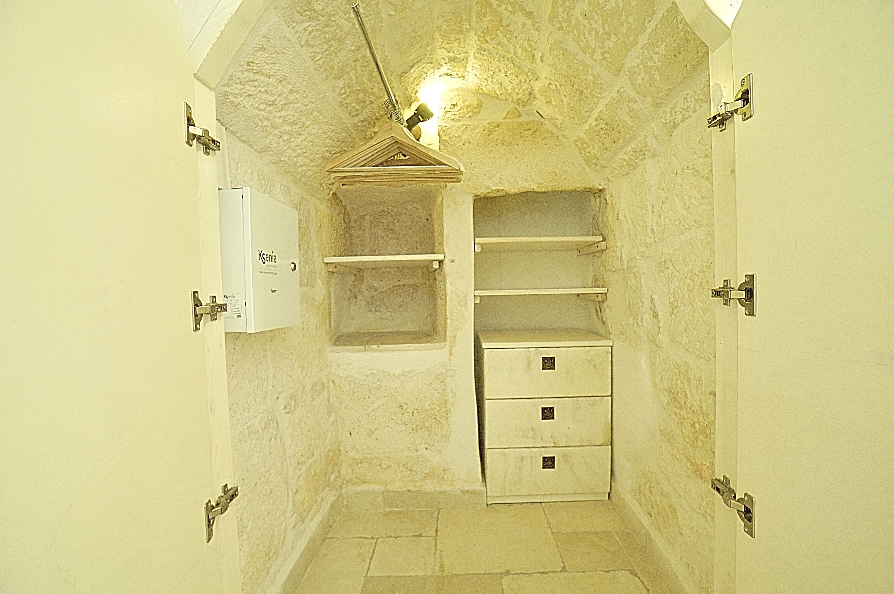 Una habitación pequeña y estrecha con una pared blanca, un estante de madera, un armario blanco y una puerta con pomo metálico.