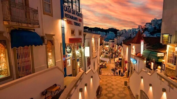 A narrow street with buildings on either side, a restaurant named 'Rasa da Fonte' with a sign indicating a grill and terrace, and a 'Steps Bar' sign on the right. - Spacious, well equipped apartment, broadband, Sky TV, with pool just outside. (Albufeira)