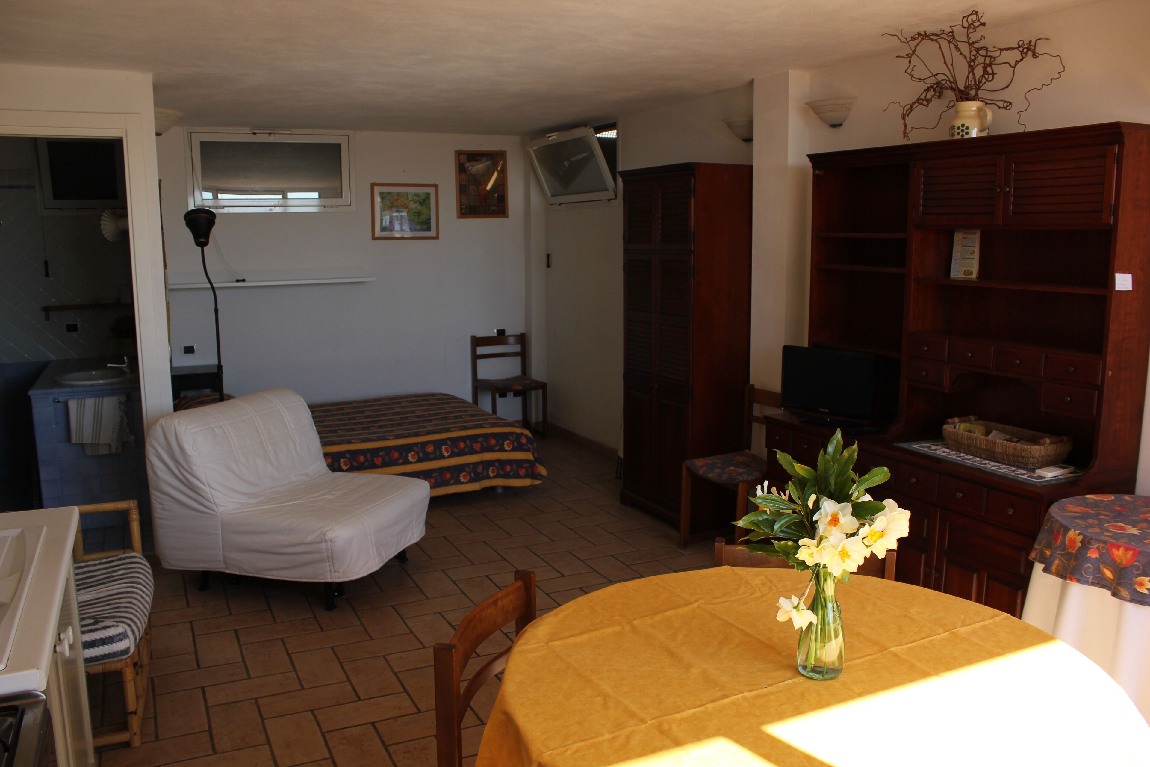 Ein kleiner, gemütlicher Raum mit einem Bett, einem Sessel, einem Tisch mit Blumenvase, einem Fernseher und einer Holzkabine.