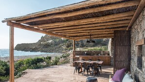 A covered outdoor seating area with a stone wall, a wooden table, and chairs, overlooking a rocky coastline. - The Old Beach House - A house by the sea to reconnect with yourself (Keramoti western crete)