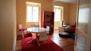 Un coin repas avec une table ronde, des chaises blanches et un tapis rouge. On y trouve également une bibliothèque rouge et une petite table ronde surmontée d’un tapis coloré.