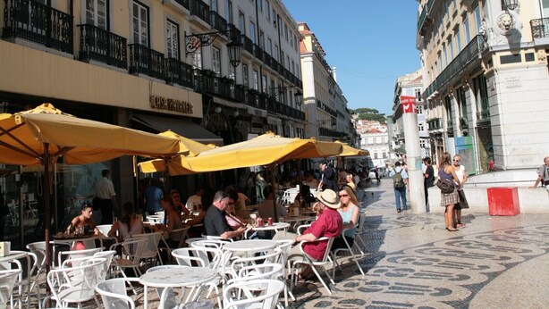 Des tables et des chaises sont disposées à l’extérieur, sur une rue pavée, et les bâtiments sont dotés de balcons et de fenêtres.