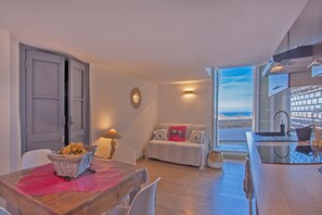 Une cuisine moderne dotée d’une grande fenêtre, un coin repas avec une table et des chaises, et un espace salon confortable avec un canapé et des coussins.