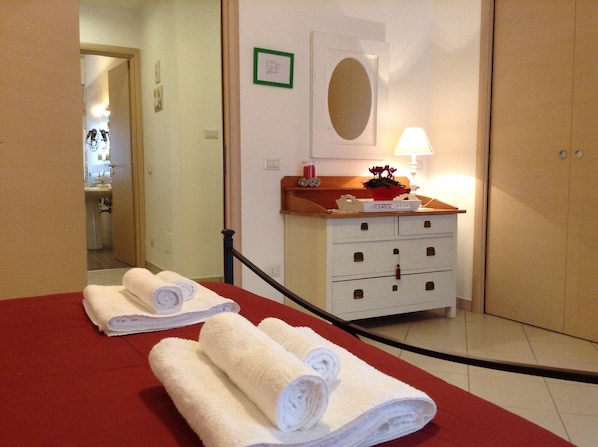 Une chambre d’hôtel avec un tapis rouge, deux serviettes blanches roulées posées sur une table, une commode en bois avec un miroir, une lampe et une petite plante en pot.