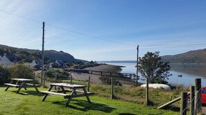 A lakeside picnic area with picnic tables, a wooden deck, and a view of houses and hills in the background. - Glenelg - Detached House With Stunning Views Over the Sea to the Isle of Skye (Glenelg)