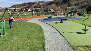 Ein Spielplatz mit Schaukeln, Rutschen und Klettergerüsten, umgeben von einer Rasenfläche und einem Weg.