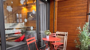 Een terras met houten vloer, een rode metalen tafel en stoelen, een raam met decoratieve sneeuwvlokken en een plant in pot.