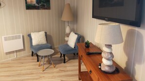 Una sala de estar con televisor, dos sillones azules, una mesita redonda, una lámpara y un cuadro en la pared.