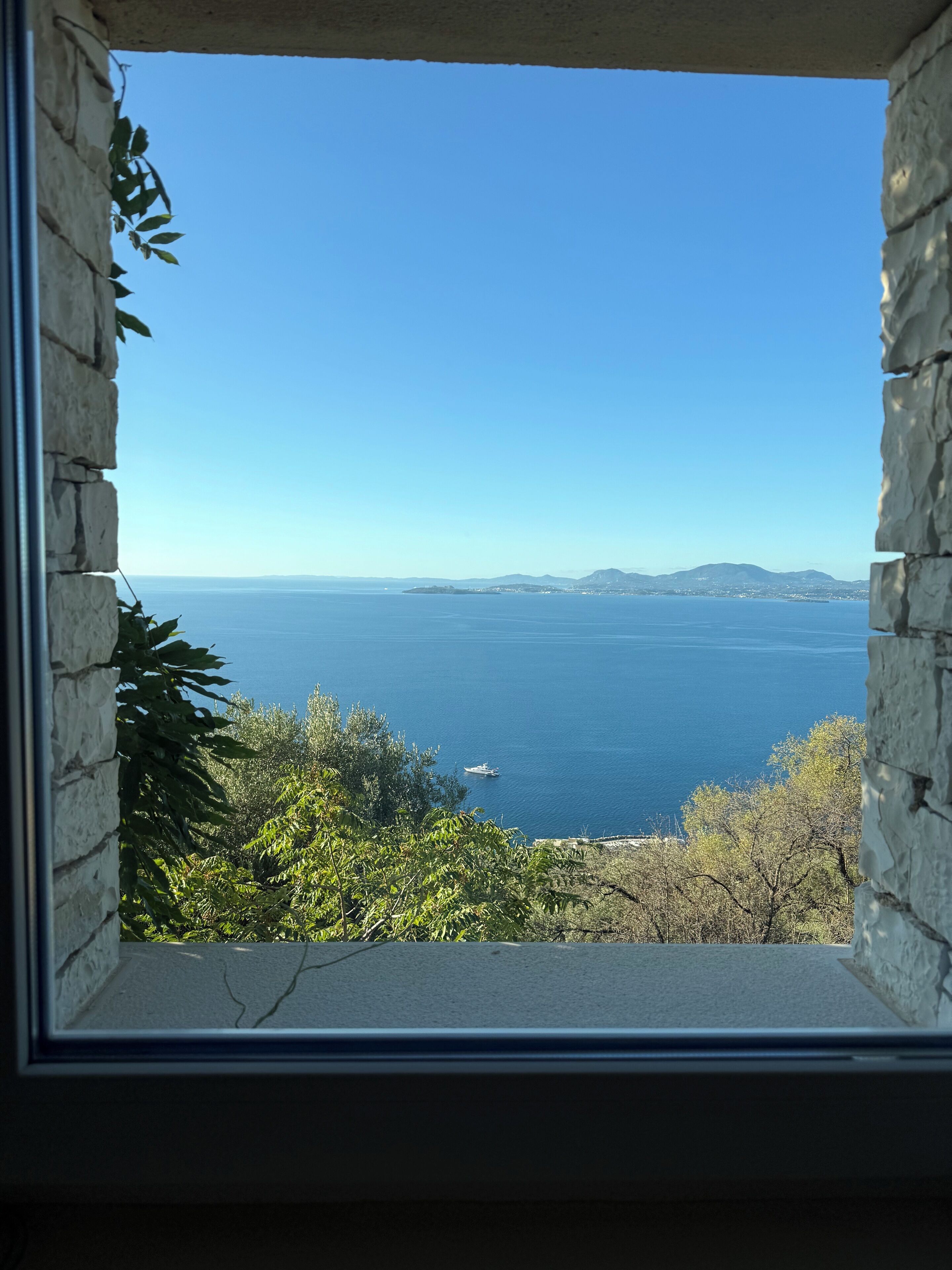 Blick durch ein steinerne Fenster, das auf eine Küstenlandschaft mit blauem Himmel und fernen Bergen blickt.