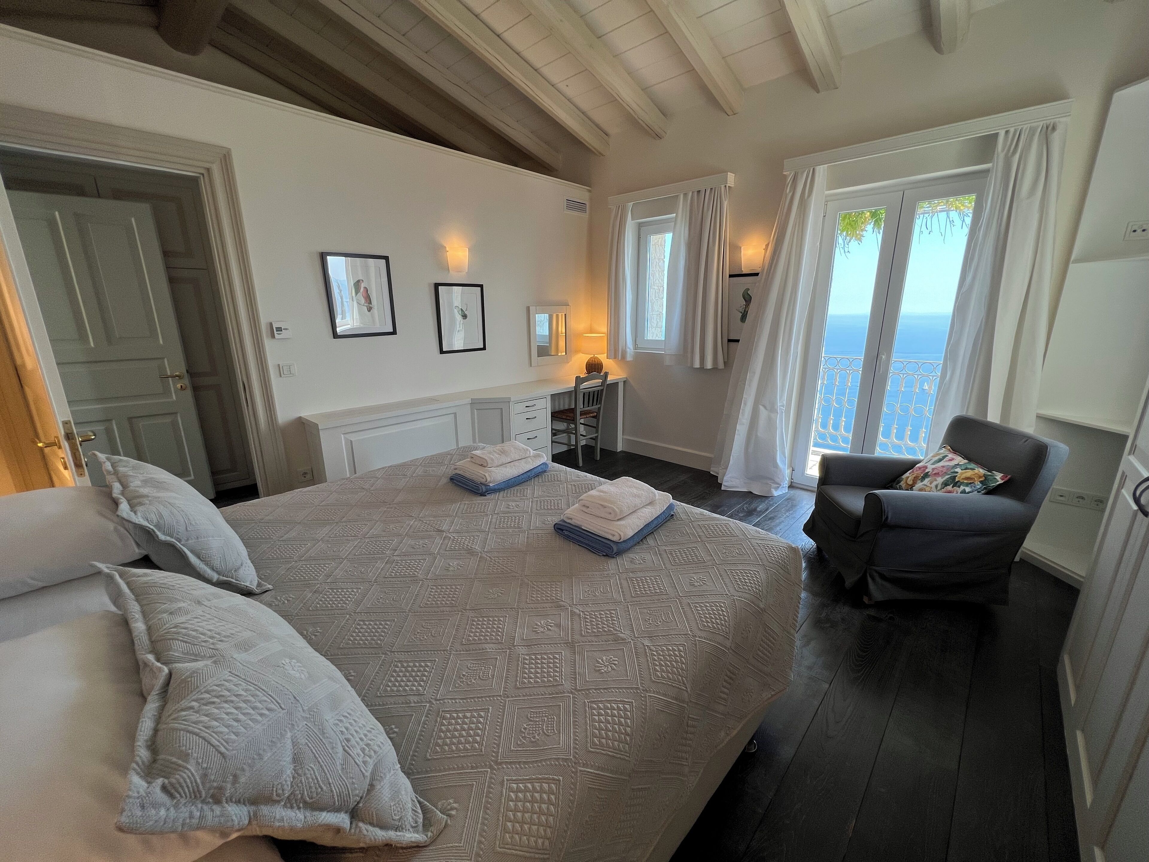 Een slaapkamer met een groot bed, een stoel, een bureau en uitzicht op zee door de ramen.