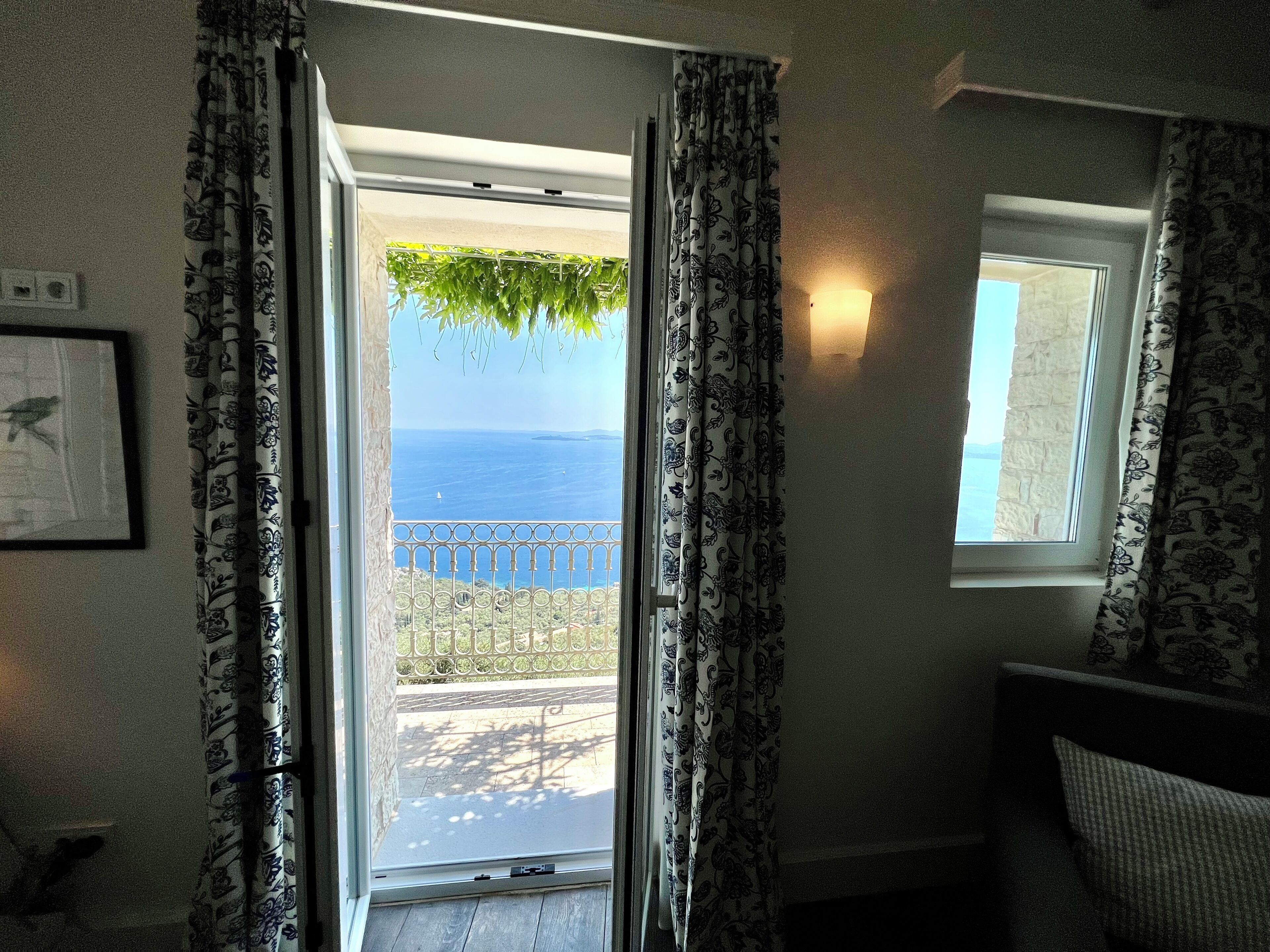 Une chambre avec vue sur la mer, accessible par une porte-fenêtre donnant sur un balcon, des rideaux à motifs, un tableau encadré au mur et une petite fenêtre offrant également une vue.