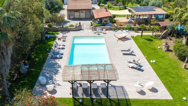 Una piscina grande con tumbonas y una zona de estar cubierta, rodeada de un jardín cuidado y una casa con techo de tejas.