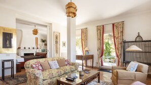 Un salon avec un canapé à motifs floraux, une table basse en bois et un fauteuil beige. On y trouve une cheminée avec une manteau de cheminée, une table d’appoint et un luminaire suspendu.