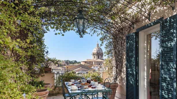 Un espace extérieur aménagé pour les repas, avec une table dressée pour deux, entouré de plantes en pot et offrant une vue sur un bâtiment historique en arrière-plan.