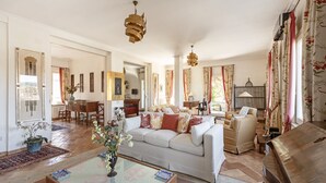 Un salon spacieux avec un canapé blanc, des fauteuils beiges et une table basse. On y trouve deux lampes suspendues et une grande fenêtre ornée de rideaux à motifs floraux.