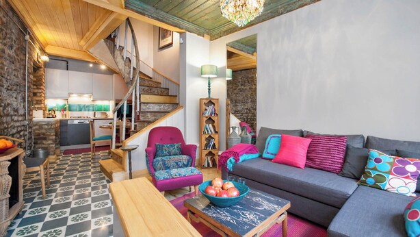 Ein gemütliches Wohnzimmer mit Schachbrettmuster-Boden, einer Holztreppe, einer Couch mit bunten Kissen, einem Bücherregal und einem Essbereich mit Tisch und Stühlen.