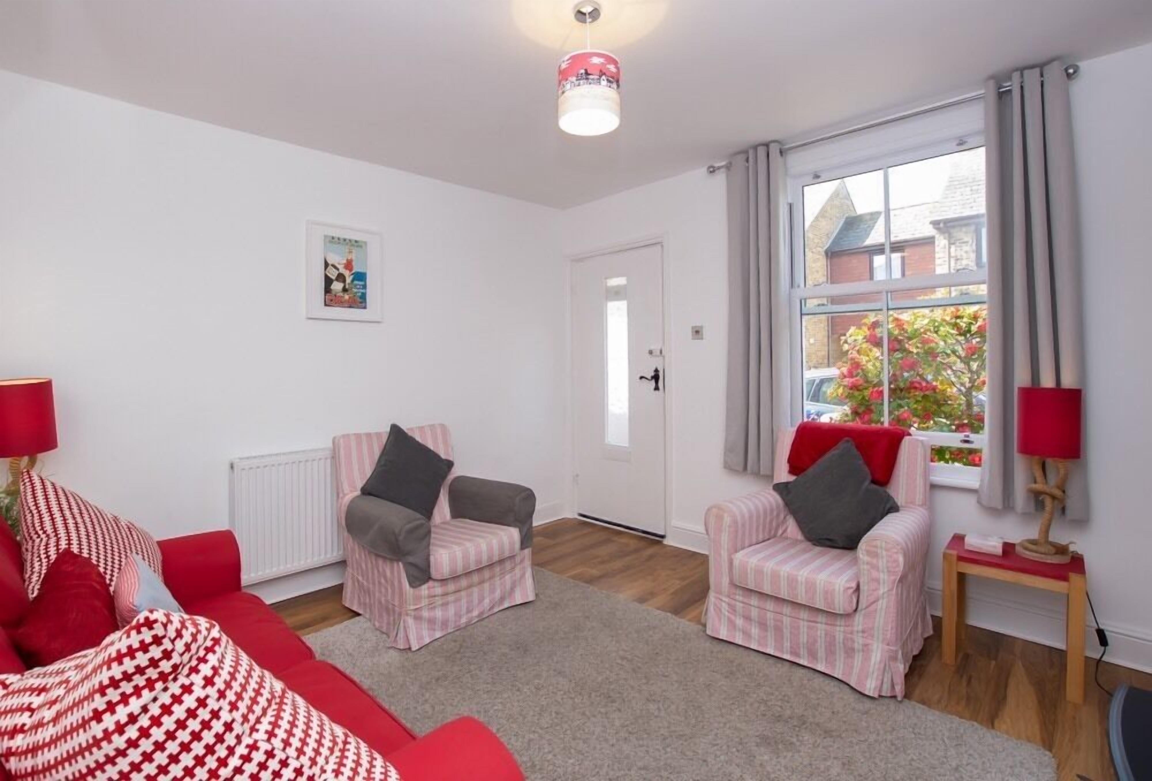 Un salon avec deux fauteuils roses, un canapé rouge et une petite table avec une lampe. Il y a une fenêtre avec des rideaux et un radiateur au mur.