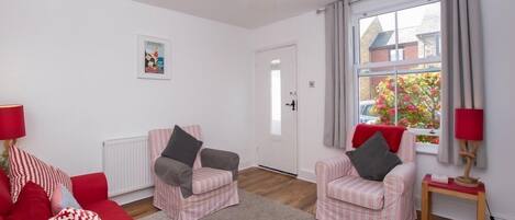 Un salon avec deux fauteuils roses, un canapé rouge et une petite table avec une lampe. Il y a une fenêtre avec des rideaux et un radiateur au mur.