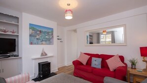 A living room with a red sofa, a television, a fireplace, and a painting of a coastal town. - Seashell Cottage - 50 meters from the beach (Walmer)