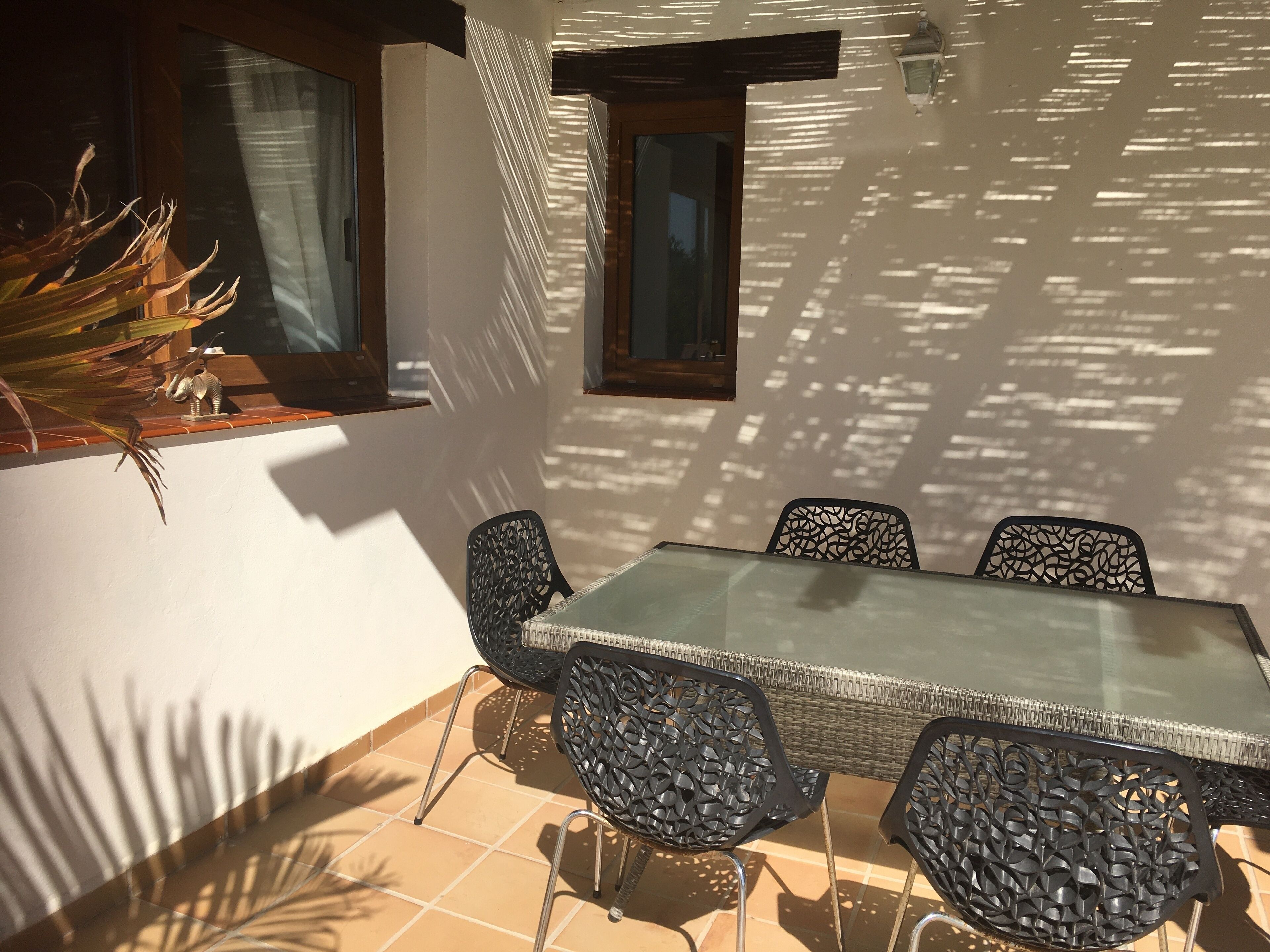 A sunlit patio with a glass table and metal chairs, a potted plant, and a window casting shadows on the wall.