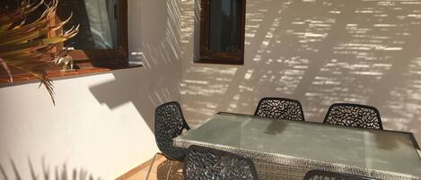 A sunlit patio with a glass table and metal chairs, a potted plant, and a window casting shadows on the wall.