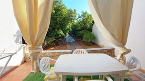 A patio with a white table and chairs, surrounded by yellow curtains, and a view of a garden with trees and a stone pathway.