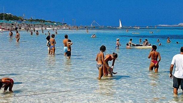 A crowded beach with people in the water and on the shore, boats in the background, and a clear blue sky.