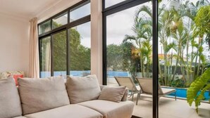 A modern living room with a large window, a beige sofa, and a view of a pool and palm trees. - Rochinha’s house with fabulous views over the bay of funchal (Funchal)