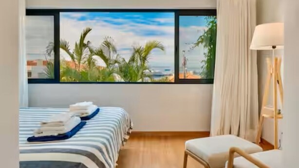 A hotel room with a large window offering a view of palm trees and the sea. There is a bed with a striped bedspread and a chair with a lamp. - Rochinha’s house with fabulous views over the bay of funchal (Funchal)