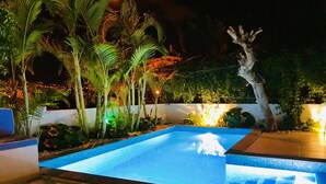 A swimming pool with a tiled edge, surrounded by palm trees and plants, with a white wall and a tree with exposed roots in the background. - Rochinha’s house with fabulous views over the bay of funchal (Funchal)